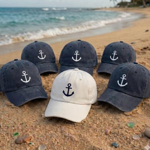 May include: Six baseball caps with an anchor design embroidered on the front. Five caps are navy blue, and one is off-white. The caps are arranged on a sandy beach with the ocean in the background.