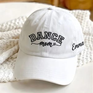 May include: A white baseball cap with the words "DANCE mom" embroidered in black script, with a small heart. The name "Emma" is also embroidered on the side. The cap is made of a soft fabric.