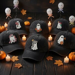 May include: Six black baseball caps arranged in a group, each featuring an embroidered patch of a white ghost adorned with various colorful flowers. The caps are displayed on a dark wooden surface alongside small pumpkins and lit candles.