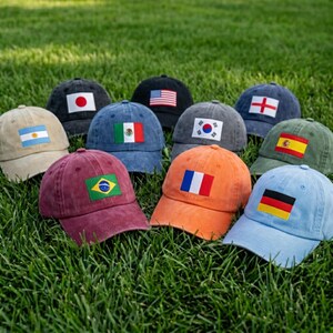 May include: A collection of baseball caps, each displaying a flag from different countries. The caps are in various colors, including black, blue, and orange, and are arranged on a bed of green grass. The flags represent countries such as the USA, Japan, and France.