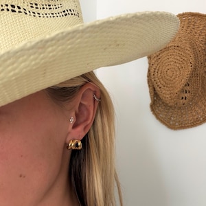 May include: A gold-toned triple-band hoop earring worn in the earlobe, featuring a polished, layered design. The ear also displays a small silver-toned cartilage hoop and a tiny, clear, marquise-cut stud earring in the upper lobe area.
