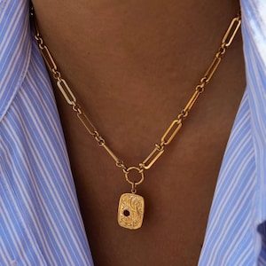 May include: Gold-coloured necklace with a rectangular pendant featuring a red gemstone. The pendant has an ornate, engraved design. The necklace is composed of a chain with elongated links and is worn against a blue and white striped shirt.