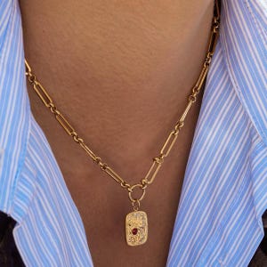 May include: A gold-toned necklace featuring a long, rectangular link chain and a rectangular pendant. The pendant has an intricate embossed floral pattern and a small, centred red gemstone. The necklace is worn over a blue and white striped shirt.