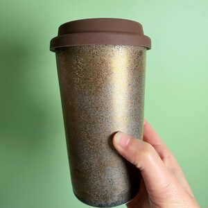 May include: A travel mug with a brown lid and a metallic gold and bronze speckled body. The reusable coffee cup is held by a hand against a light green background. The travel mug is designed to keep beverages hot or cold.
