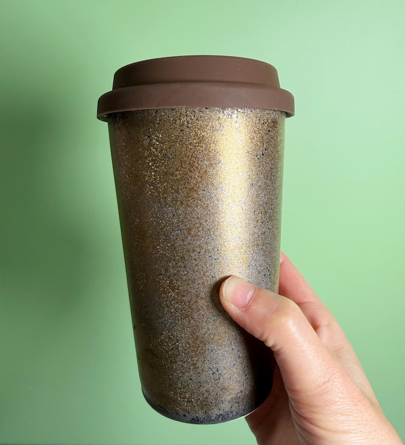 May include: A travel mug with a brown lid and a metallic gold and bronze speckled body. The reusable coffee cup is held by a hand against a light green background. The travel mug is designed to keep beverages hot or cold.