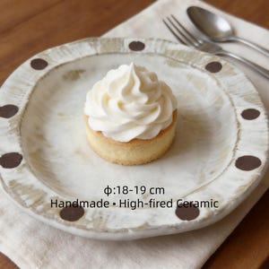 May include: A handmade, high-fired ceramic plate with a diameter of 7-7.5 inches, featuring a white glaze and brown dot accents. A small cake topped with white frosting sits on the plate. The plate is on a linen napkin.