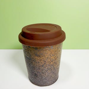 May include: A reusable coffee cup with a brown silicone lid. The cup has a speckled design in shades of brown, black, and gold. The cup is designed to be used for hot or cold beverages.