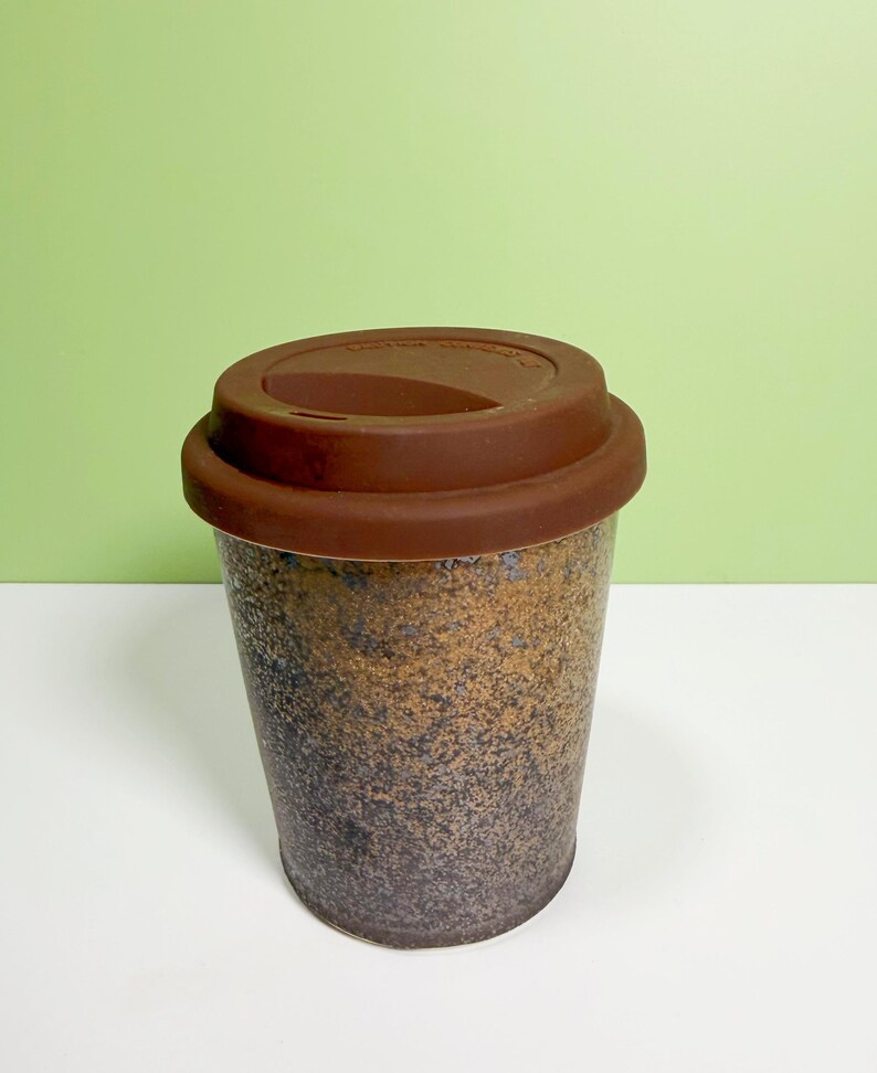May include: A reusable coffee cup with a brown silicone lid. The cup has a speckled design in shades of brown, black, and gold. The cup is designed to be used for hot or cold beverages.