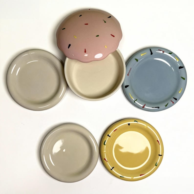 May include: A set of ceramic plates and a serving dish. The plates are in various colors, including beige, blue, and yellow, with colorful speckles around the rims. The serving dish has a pink lid with colorful sprinkles.
