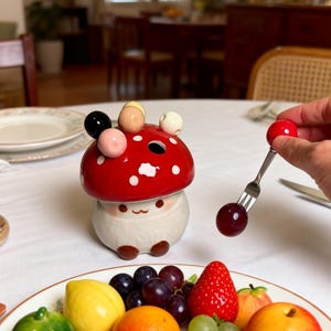 May include: A novelty mushroom-shaped ceramic condiment holder with a red cap and white spots. The holder has a smiling face and is next to a plate of fresh fruit, including grapes, strawberries, and lemons. A person is holding a red cherry on a fork.