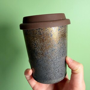 May include: A ceramic travel mug with a brown silicone lid. The mug has a speckled, textured surface with a gradient of colors, transitioning from black at the base to gold and brown at the top. The mug is held by a hand.