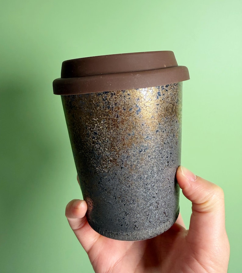 May include: A ceramic travel mug with a brown silicone lid. The mug has a speckled, textured surface with a gradient of colors, transitioning from black at the base to gold and brown at the top. The mug is held by a hand.