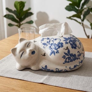 Może przedstawiać: Białe ceramiczne pudełko na chusteczki w kształcie śpiącego kota. Kot ma niebieskie akcenty na pyszczku i wokół oczu. Ciało zdobią niebieskie wzory kwiatowe. Białe chusteczki wychodzą z grzbietu kota.