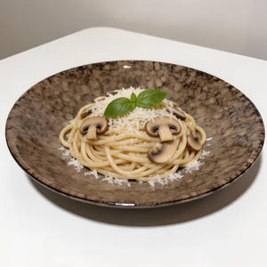 Puede incluir: Plato de espaguetis con champiñones en rodajas, queso rallado y una guarnición de hoja de albahaca. Servido en un cuenco de cerámica marrón moteado. El plato se presenta sobre una superficie blanca.