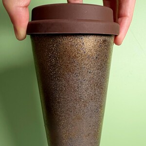 May include: A reusable coffee cup with a brown silicone lid. The cup has a speckled, metallic bronze finish. The cup is held by a person's hand against a light green background. The cup is designed for hot or cold beverages.
