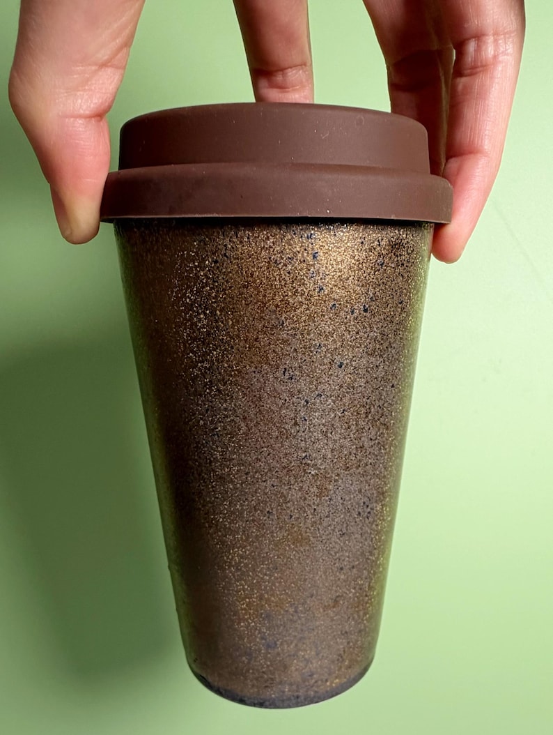 May include: A reusable coffee cup with a brown silicone lid. The cup has a speckled, metallic bronze finish. The cup is held by a person's hand against a light green background. The cup is designed for hot or cold beverages.