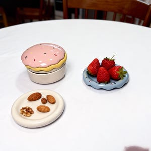 May include: A collection of ceramic serving dishes and a decorative container. The container is shaped like a donut with pink icing and sprinkles. A small blue plate holds strawberries, and a white plate holds nuts.