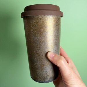 May include: A reusable travel coffee mug with a brown lid and a textured gold and bronze exterior. The mug is designed to keep beverages hot or cold and is held by a hand against a green background.