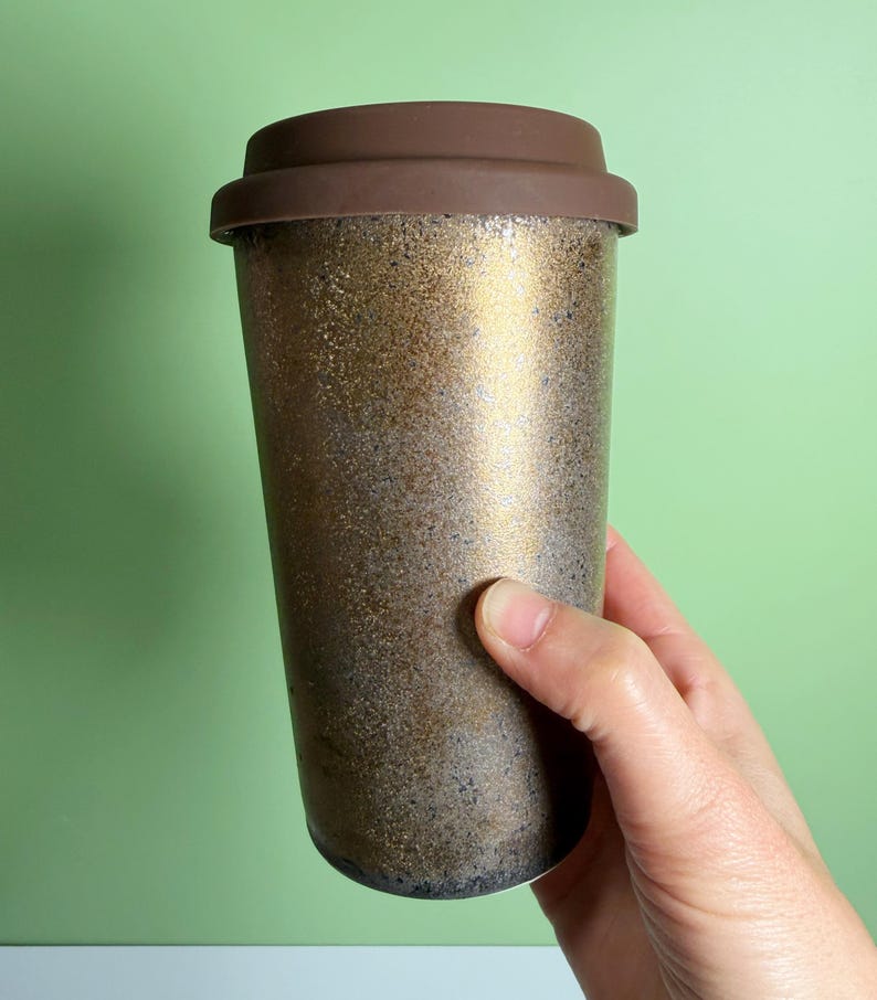 May include: A reusable travel coffee mug with a brown lid and a textured gold and bronze exterior. The mug is designed to keep beverages hot or cold and is held by a hand against a green background.