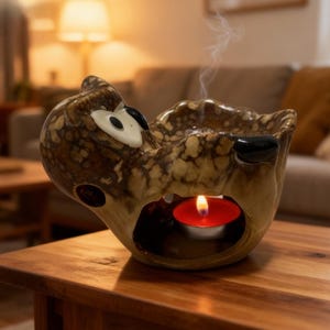 Può includere: Un diffusore di oli essenziali a forma di gufo in ceramica con una candela tealight accesa all'interno. Il gufo è marrone e beige con dettagli neri e bianchi. Il fumo sale dalla parte superiore, indicando la diffusione di oli essenziali. Il diffusore è su un tavolo di legno.