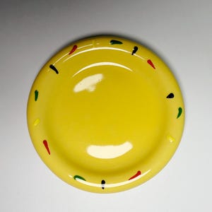 May include: A bright yellow ceramic plate with a slightly raised rim. The plate is decorated with small, colorful dashes of red, green, yellow, and black around the edge. The plate has a glossy finish.