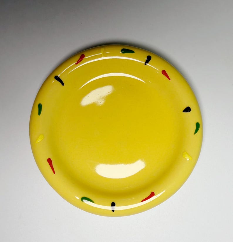 May include: A bright yellow ceramic plate with a slightly raised rim. The plate is decorated with small, colorful dashes of red, green, yellow, and black around the edge. The plate has a glossy finish.