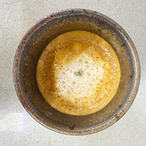 May include: Overhead view of a ceramic bowl filled with a frothy, light brown liquid. The liquid has a foamy, bubbly texture with a darker center. The bowl is a dark, earthy brown color with a textured rim, set against a light-colored surface.