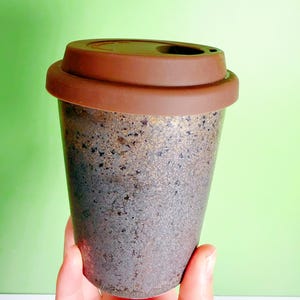 May include: A reusable coffee cup with a brown silicone lid. The cup has a speckled, textured surface with shades of gray, brown, and gold. The lid has a small opening for drinking. The cup is designed for hot or cold beverages.