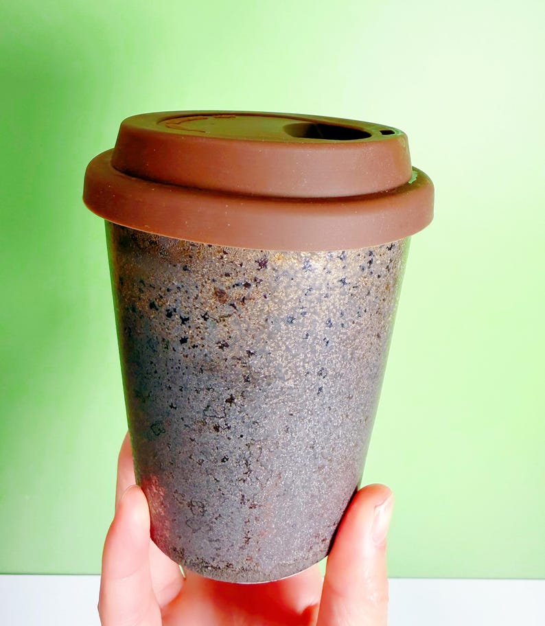 May include: A reusable coffee cup with a brown silicone lid. The cup has a speckled, textured surface with shades of gray, brown, and gold. The lid has a small opening for drinking. The cup is designed for hot or cold beverages.