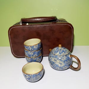 Może przedstawiać: Zestaw do herbaty w stylu vintage z brązową skórzaną walizką. Zestaw zawiera czajniczek i cztery filiżanki, wszystkie zdobione niebiesko-białym kwiatowym wzorem. Czajniczek ma brązowy uchwyt i drewnianą gałkę na pokrywce.
