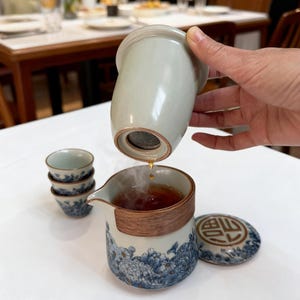 May include: A tea set with a light green infuser, pouring tea into a white and blue floral patterned teapot. The set includes small matching teacups and a lid with a Chinese character design. The teapot has a wooden band around the middle.