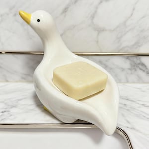 Op de afbeelding: Een witte keramische zeepbak in de vorm van een eend, met een gele snavel en poten. Een vierkante zeep van lichtbeige zeep ligt in de bak. De achtergrond is een marmerpatroon.
