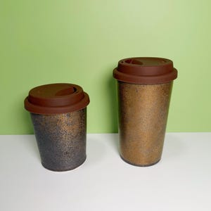 Może przedstawiać: Dwa kubki podróżne z brązowymi pokrywkami. Kubki mają nakrapiane metaliczne wykończenie w odcieniach brązu i czerni. Jeden kubek jest krótszy i szerszy od drugiego. Pokrywki wykonane są z ciemnobrązowej gumy. Tło jest jednolite, jasnozielone.