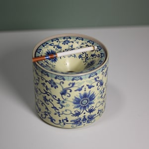May include: A cylindrical ceramic ashtray with a floral design in blue on a cream background. A lit cigarette rests in the ashtray. The ashtray has a circular opening and a slightly raised rim. The design is reminiscent of traditional Chinese porcelain.