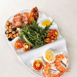 May include: A white divided serving platter containing a meal with bacon-wrapped items, roasted sweet potato, chickpeas, black beans, fresh green frisée lettuce, halved cherry tomatoes, a small pool of oil, hard-boiled egg halves, and shrimp.