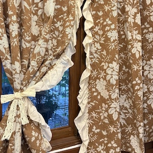 Puede incluir: Cortinas florales marrones con estampados de flores blancas. Las cortinas tienen un borde con volantes color crema y están atadas con un lazo de encaje color crema. Las cortinas cuelgan frente a un marco de ventana de madera.