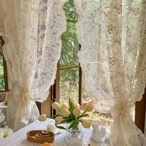 May include: Delicate cream lace curtains adorn a window, with a table set for a meal. A vase of yellow tulips sits near a plate of food and a cup of coffee. The curtains are tied back with matching fabric.