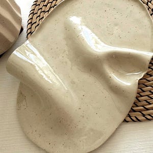 May include: A beige ceramic plate with a distinctive, undulating design. The plate features a speckled surface and a glossy finish. It rests on a woven placemat. Plate dimensions are not visible.