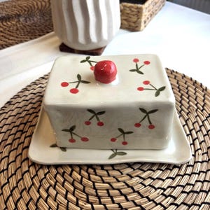May include: A ceramic butter dish with a lid and a matching rectangular base. The dish is off-white with a pattern of red cherries and green leaves. A single red cherry serves as a handle on the lid. The dish sits on a woven placemat.