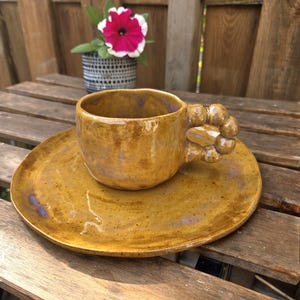 Op de afbeelding: Een handgemaakte keramische kop en schotel set in een warme, aardse tint. De kop heeft een uniek handvatontwerp en staat op een bijpassende schotel. De set staat op een houten oppervlak, met een bloempot op de achtergrond.
