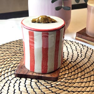 Peut inclure: Un pot en céramique avec un couvercle orné d'une poignée en forme de croissant. Le pot présente un motif à rayures rouges et blanches et repose sur un petit dessous de verre en bois. L'arrière-plan comprend un vase rose et un set de table tissé.