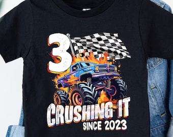 May include: Black t-shirt with a monster truck graphic, the number 3, and a chequered flag. The design includes the text "Crushing It Since 2023" and flames. Bold typography and vibrant colours.