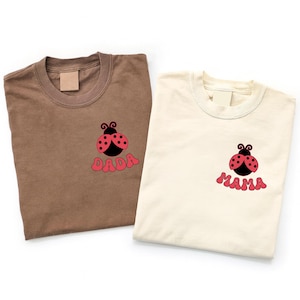 May include: Two t-shirts, one brown and one cream, each with a ladybug graphic. The brown shirt has the word "DADA" in pink, while the cream shirt says "MAMA". Both shirts are folded.