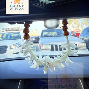 Peut inclure: Un d&eacute;sodorisant de voiture floral blanc suspendu au r&eacute;troviseur. Le d&eacute;sodorisant est compos&eacute; de fleurs blanches et de perles en bois. Le texte "ISLAND CLAY CO." est visible.