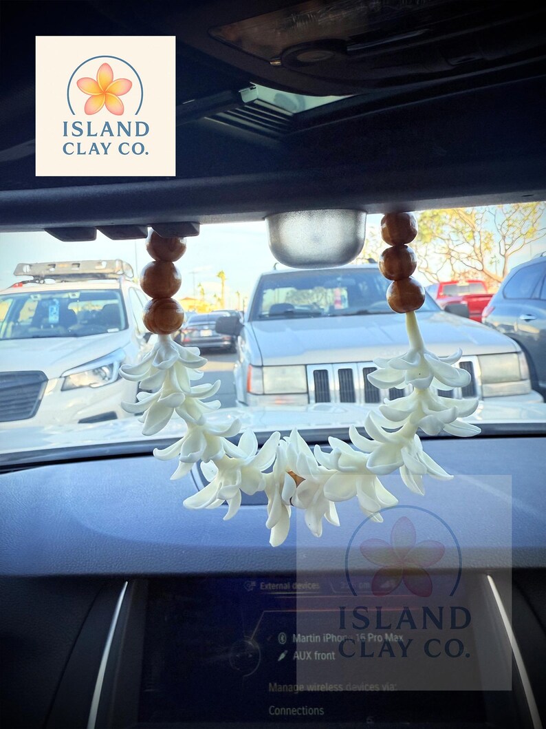 Peut inclure: Un d&eacute;sodorisant de voiture floral blanc suspendu au r&eacute;troviseur. Le d&eacute;sodorisant est compos&eacute; de fleurs blanches et de perles en bois. Le texte "ISLAND CLAY CO." est visible.