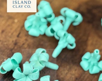 May include: Close-up of several small, light teal-colored clay flower shapes. The flowers are scattered on a wooden surface. The Island Clay Co. logo is visible in the upper left corner.