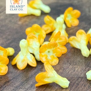 May include: Close-up of several small, yellow and orange flower-shaped clay beads. The beads have a three-petal design with a yellow stem and orange petals. The logo for Island Clay Co. is in the upper left corner.