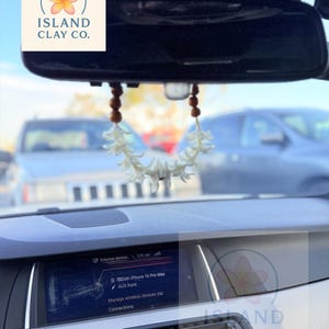 Peut inclure: Int&eacute;rieur de voiture avec r&eacute;troviseur orn&eacute; d'un lei floral et de perles en bois. L'&eacute;cran du tableau de bord affiche des informations sur les appareils externes et les connexions sans fil. Le logo Island Clay Co. est visible.