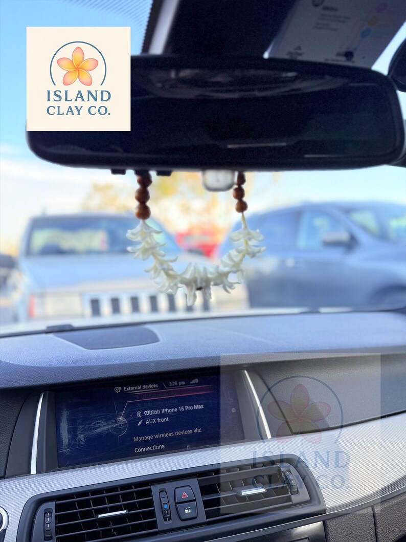 Peut inclure: Int&eacute;rieur de voiture avec r&eacute;troviseur orn&eacute; d'un lei floral et de perles en bois. L'&eacute;cran du tableau de bord affiche des informations sur les appareils externes et les connexions sans fil. Le logo Island Clay Co. est visible.
