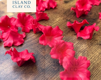 Könnte beinhalten: Leuchtend rote Tonblumen auf einer Holzoberfläche verstreut. Die Blüten befinden sich in verschiedenen Stadien der Blüte, mit detaillierten Blütenblattstrukturen. Das Logo von Island Clay Co. ist oben links sichtbar.